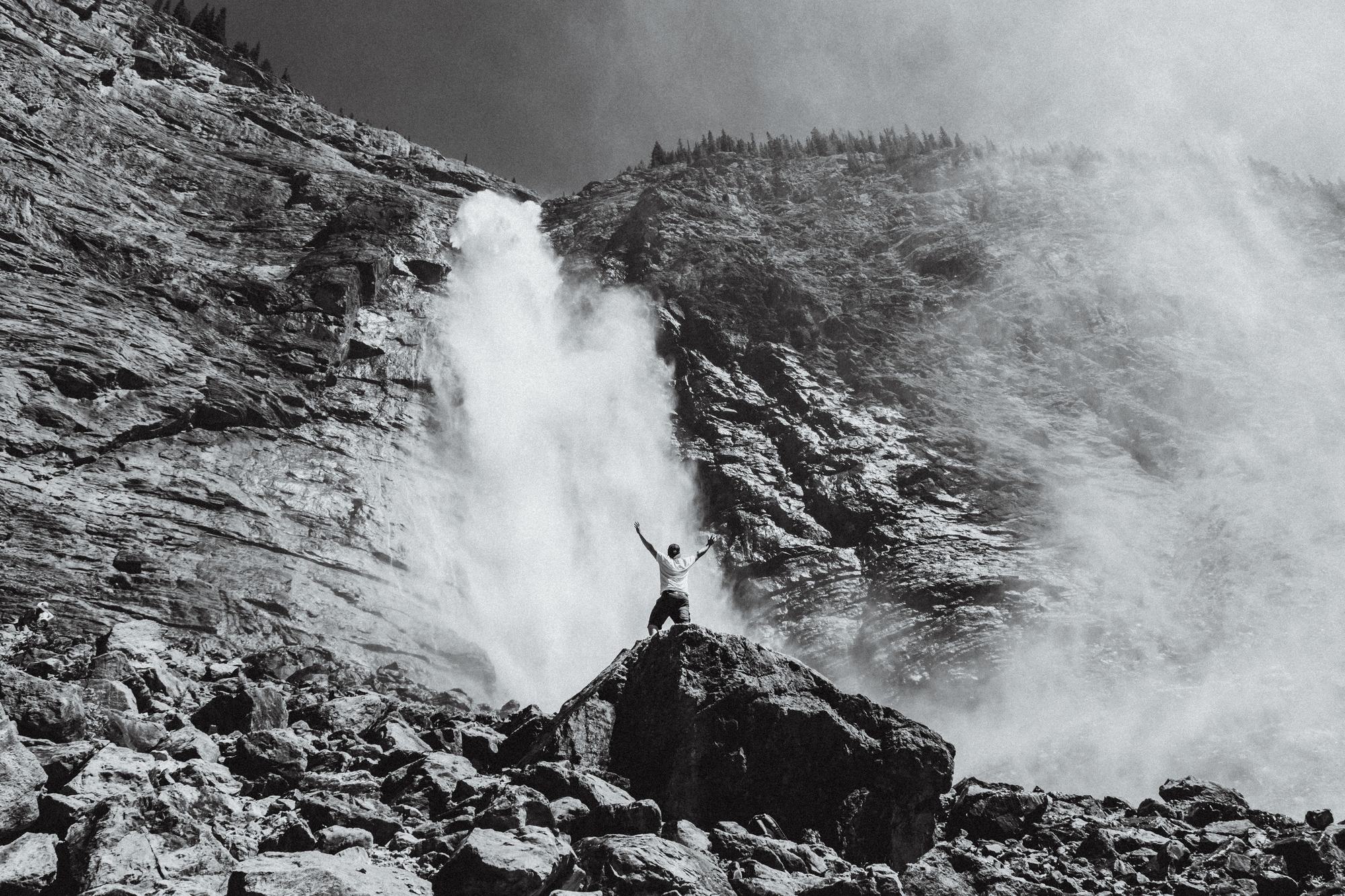 A man with raised arms on a boulder in front of a tall waterfall