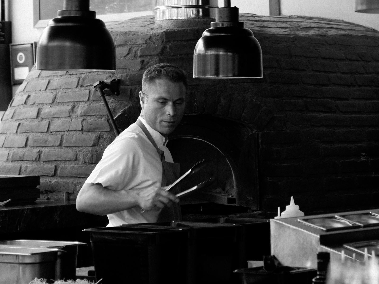 A chef in a white shirt holding tongs beside a brick oven